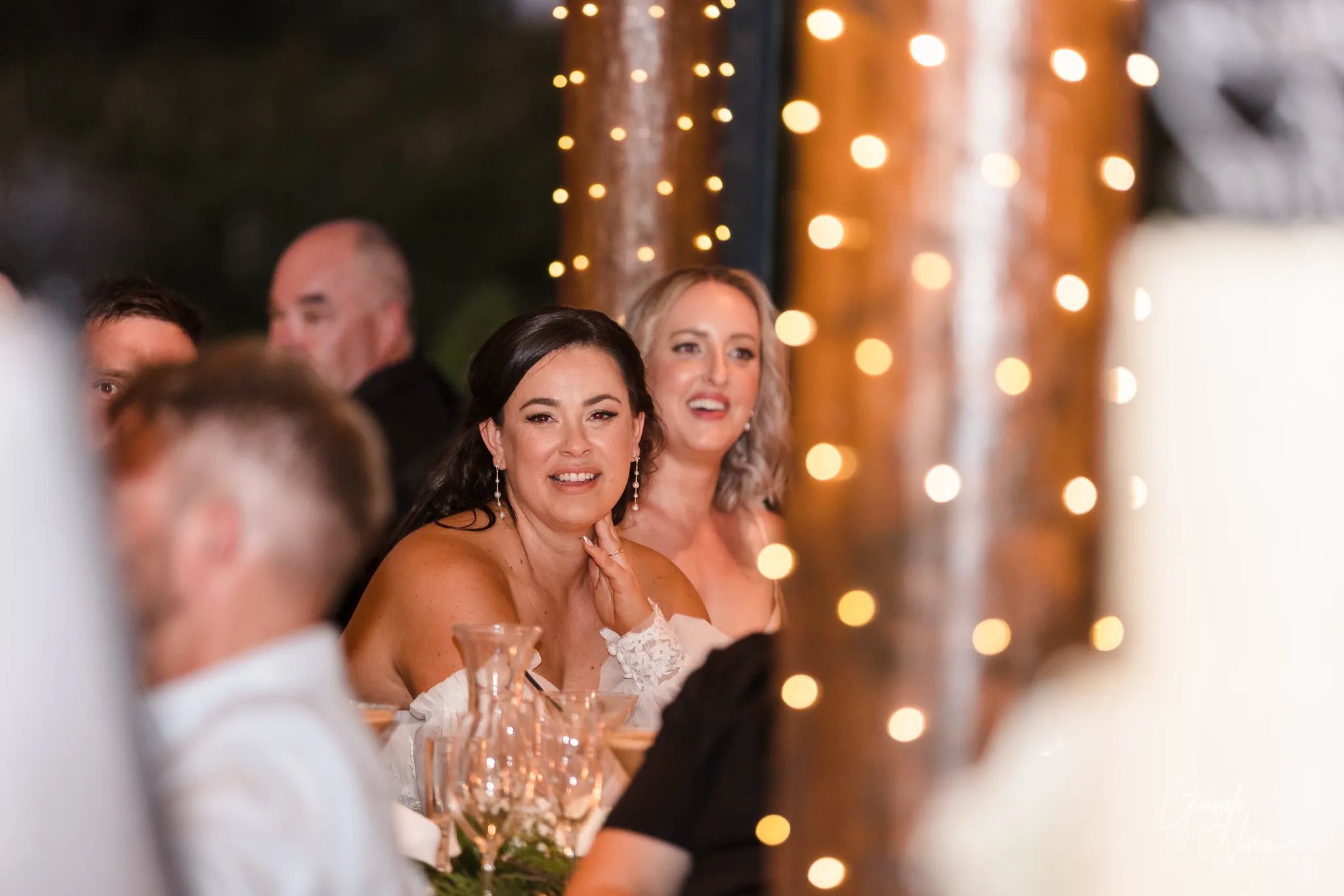 The bride and a bridesmaid are seated at a table, smiling warmly as they listen to speeches during the reception. Fairy lights softly illuminate the scene, adding a magical atmosphere to the evening.