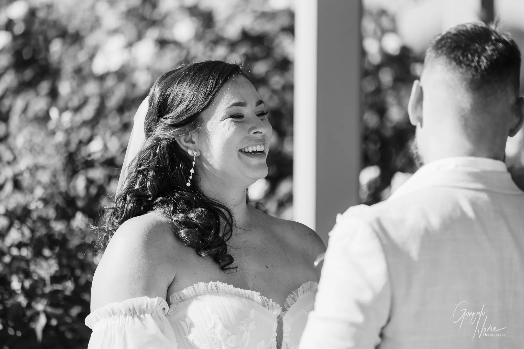 Black and white photo of bride Tahnee smiling warmly at groom James during their wedding ceremony, framed by a backdrop of soft, sunlit foliage at Sittella Winery.