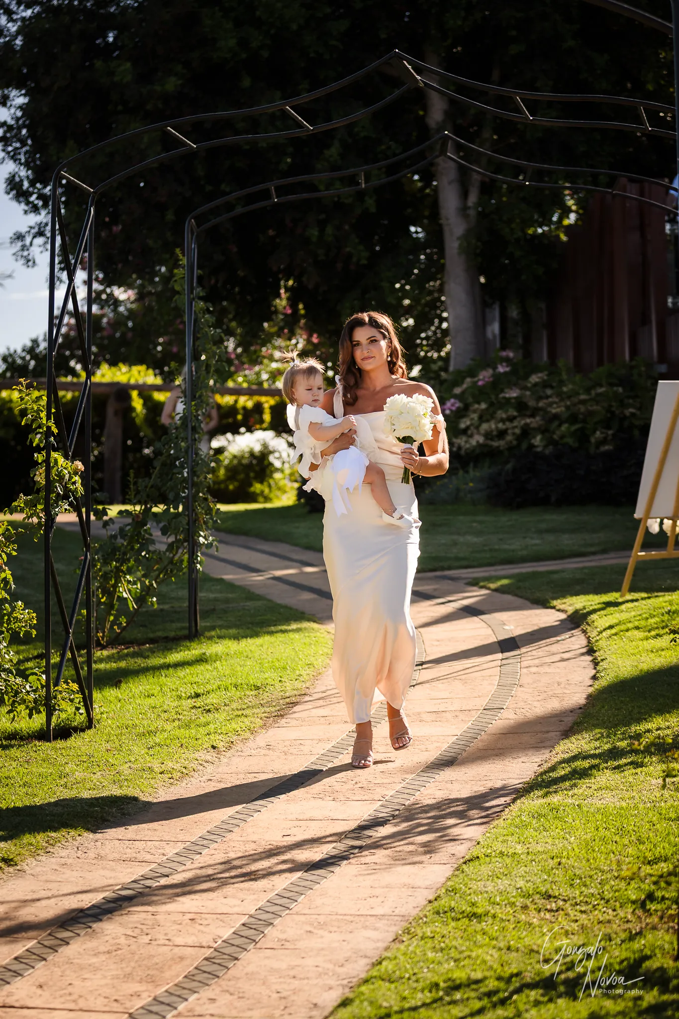 Samara in a champagne satin dress walks down the aisle at Sittella Winery holding a young flower girl and a bouquet of white flowers, both bathed in golden afternoon light.