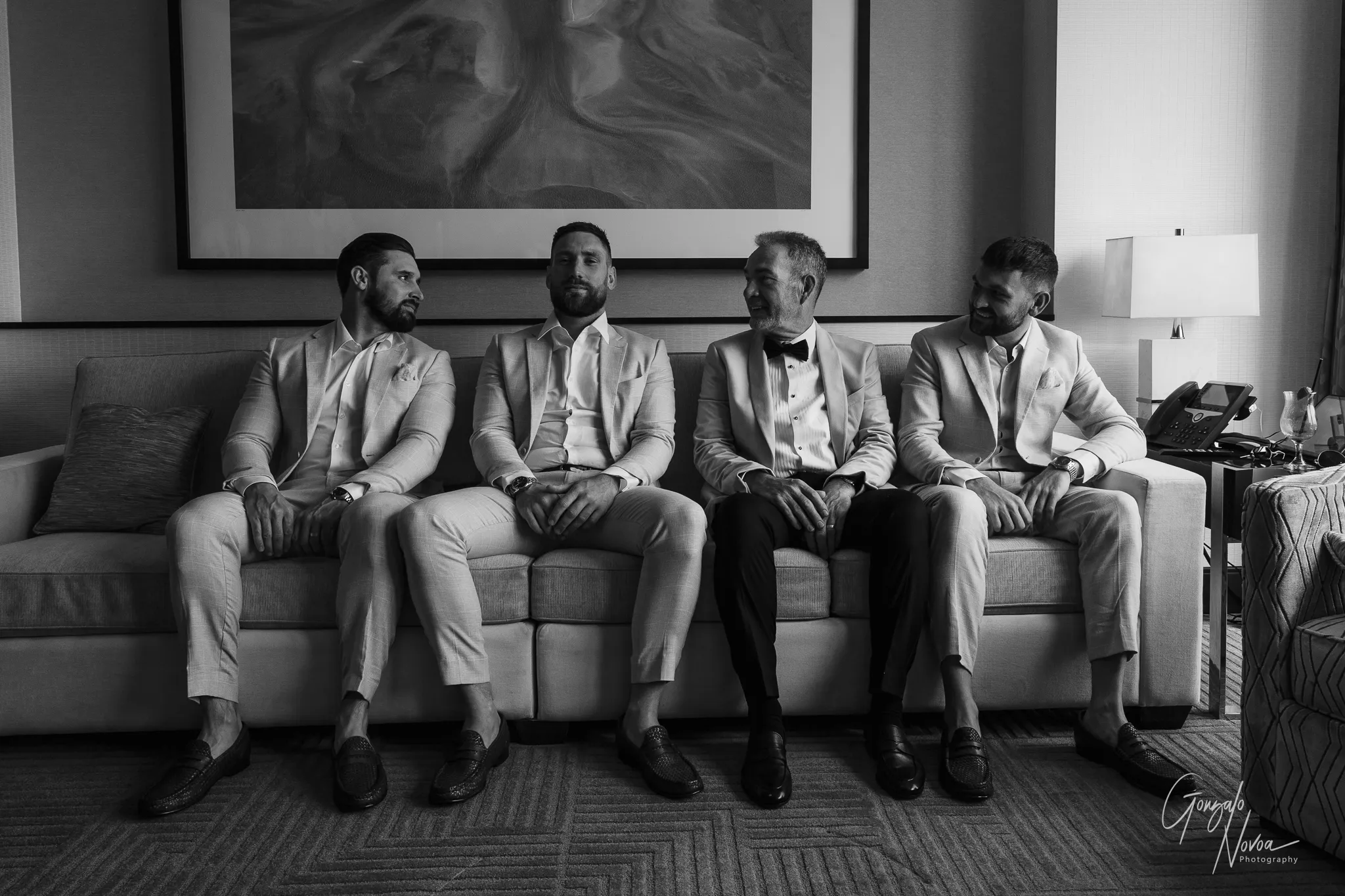 Black and white photo of James seated in the centre of a group of groomsmen and his father inside a Crown Towers Perth hotel room. Everyone is dressed in coordinated suits, smiling and relaxed, with a city skyline faintly visible through the tall windows behind them.