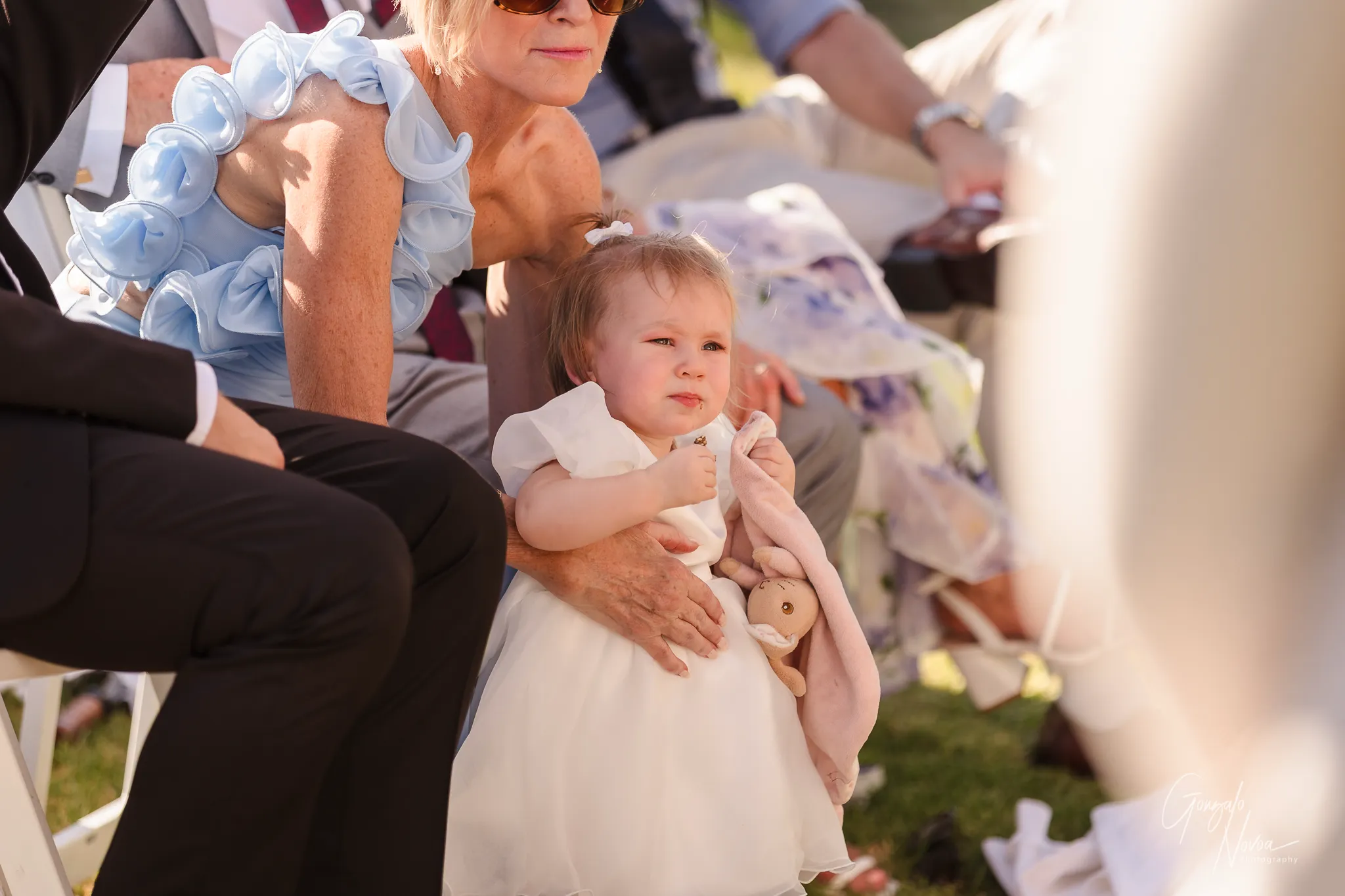 Tahnee and James’ young daughter Arlea, dressed in a white dress and holding her comfort toy, sits on a guest’s lap during the ceremony, watching her parents with a sweet and thoughtful expression.