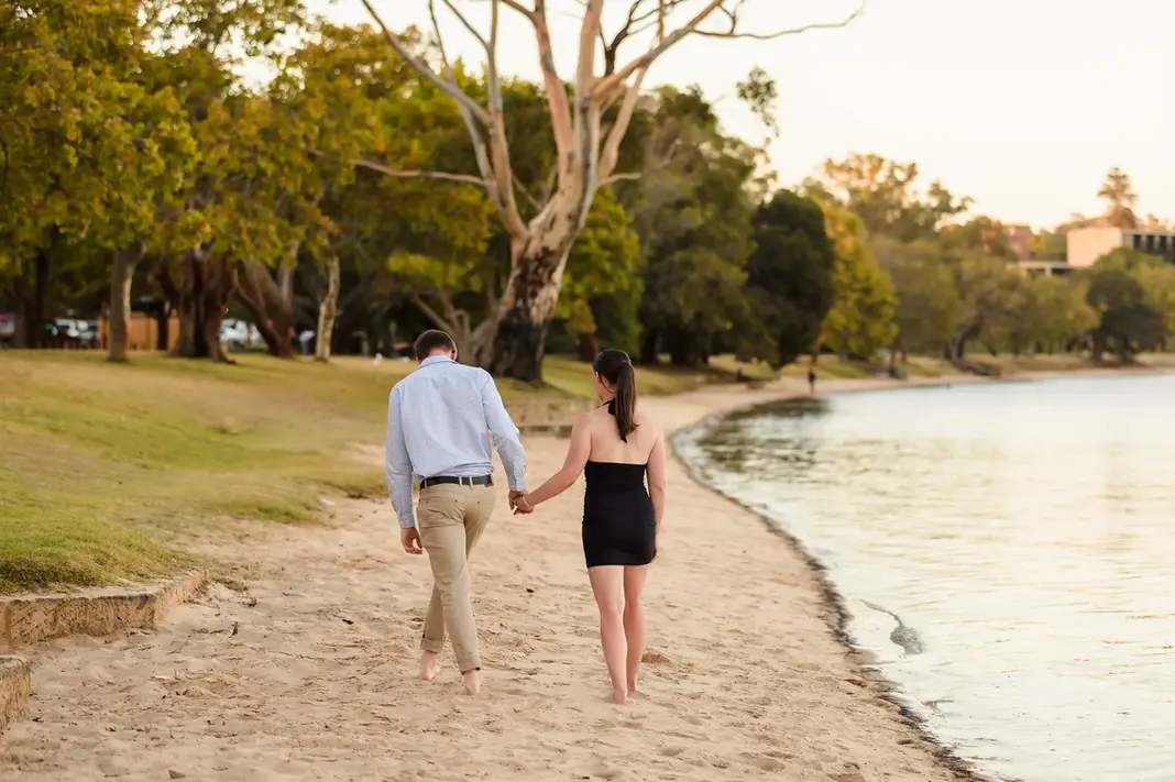 Baleigh and Kailym walking barefoot along the sandy shore at Matilda Bay Reserve, holding hands after their engagement.