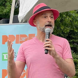 Glenn Tkach speaking into a microphone on stage at the Vancouver Pride Society Eastside Pride event