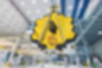 Scientists in clean suits work on the large, gold hexagonal James Webb Space Telescope in a high-tech lab with bright lighting.