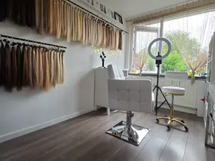 Kapsalon met witte stoel, ringlamp en haarextensies aan de muur. Grote ramen met gordijnen, donkerhouten vloer, serene sfeer.