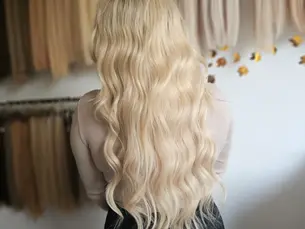 Kobieta z długimi falującymi blond włosami stoi tyłem w salonie fryzjerskim, na tle wiszących pasm włosów w różnych odcieniach.
