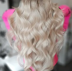 Golvend blond haar op roze trui in een onscherpe binnenruimte. De focus ligt op het haar, dat glanzend en verzorgd is.
