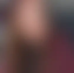 Kobieta z długimi brązowymi włosami i niebieskimi oczami w bordowej marynarce patrzy w aparat. W tle widoczna TV.