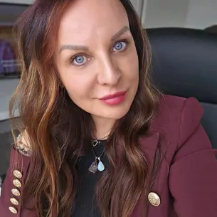 Kobieta z długimi brązowymi włosami i niebieskimi oczami w bordowej marynarce patrzy w aparat. W tle widoczna TV.