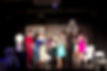An ethnically diverse group of women smile and hug each other on stage. They are wearing clothing in different colors which contrast strongly with the set on stage, which is mostly black and white and emulates a dining room and living room.