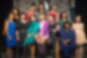 A diverse group of women stand together and smile. Behind them are decorations suggesting a hyperfeminine home including a white ornate mirror and floral drapes. They are wearing clothes in different vibrant colors.