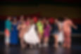 A diverse group of woman stand together and smile playfully. They are wearing vibrant clothes. Behind them are colorful swathes of fabric -- yellow, orange and blue -- lining the wall.