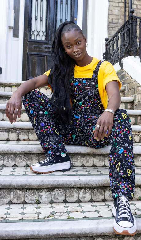 A Black woman with a long ponytail, wearing a yellow t-shirt and dungarees patterned with science-inspired motifs
