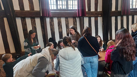 Participants of all ages cluster around two tables inside the Guildhall Museum’s timber-framed hall, working together on traditional Eastern European crafts such as doll-making and textile decoration. A workshop leader guides the group while others assist children and families during the hands-on activity.