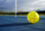 Yellow pickleball on a blue court near a net. The ball is in focus, and the court is sunlit, giving a bright and calm atmosphere.