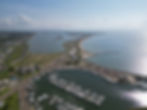Aerial view of a marina with many boats docked, roads weaving through green land, and a coastline under a blue sky with scattered clouds.