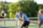 Four people playing pickleball on an outdoor court, smiling and engaged in the game. Greenery surrounds the court, creating a lively scene.