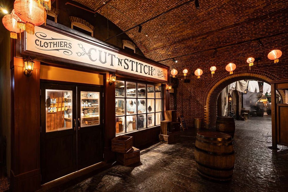 Vintage brick arcade with warm lanterns and barrels; shop sign reads CUT 'N' STITCH CLOTHIERS.