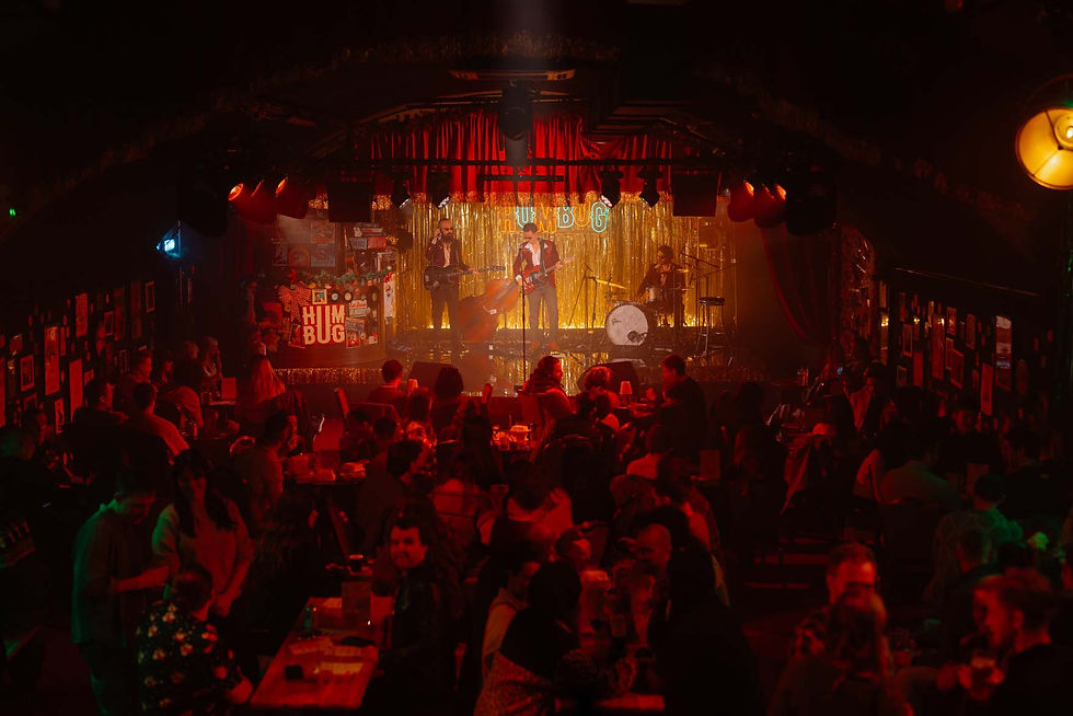 Band performs on stage with gold curtains, red lighting, and "HUMBUG" sign. Audience seated at tables, creating a lively, warm atmosphere.