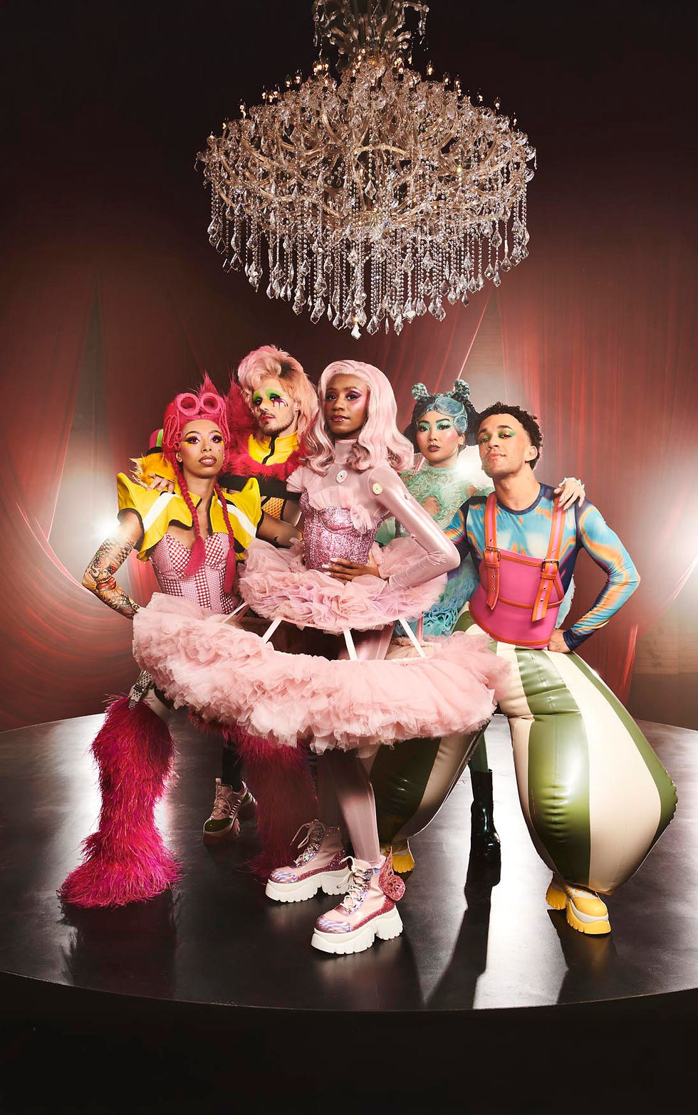 Five people in vibrant, avant-garde outfits under a chandelier, posing confidently on a stage with red curtains, exuding a bold, theatrical vibe.