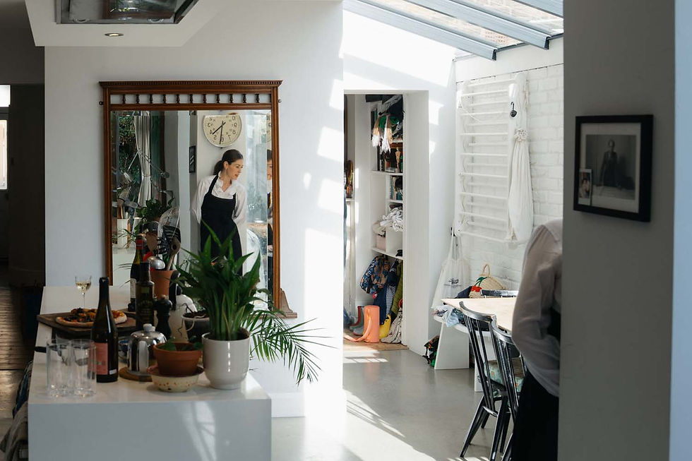 Bright home interior with a woman reflected in a mirror, dining table set with bottles and plants, under a skylit ceiling.