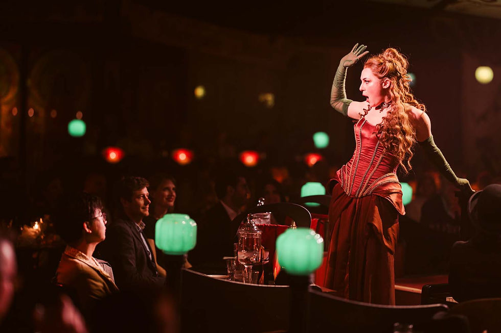 Performer in red corset poses dramatically on stage with green lamps, audience watching intently. Dimly lit, theatrical atmosphere.