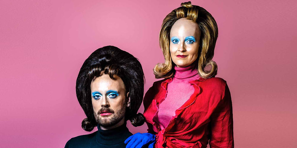 Two glam people in retro wigs and blue eye makeup pose against a pink backdrop, one in a red outfit with blue gloves.