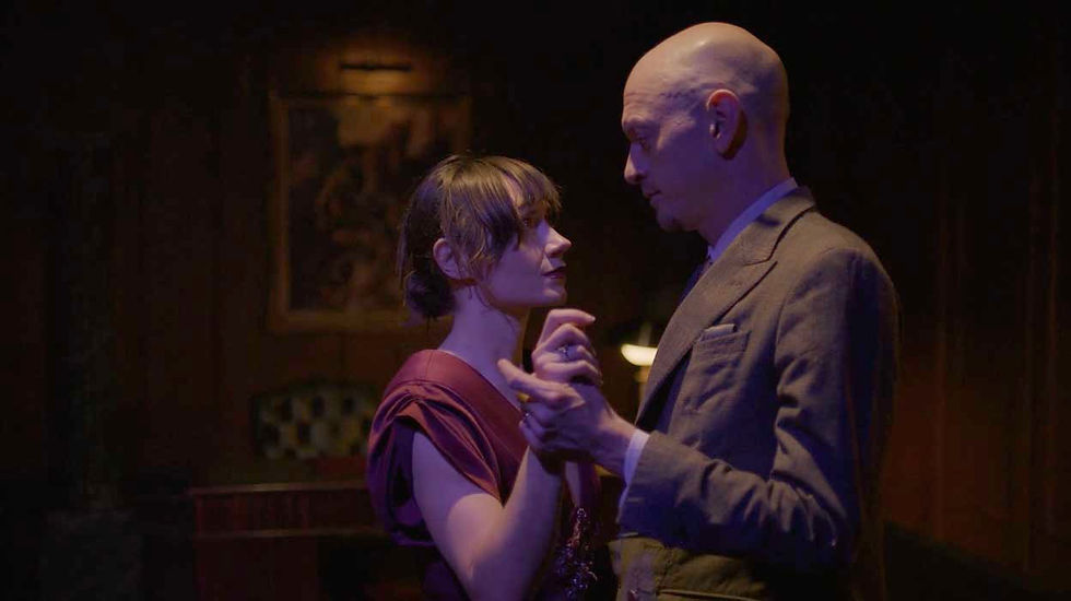 A woman in a red dress and a bald man in a suit dance closely in a dimly lit room, gazing at each other.