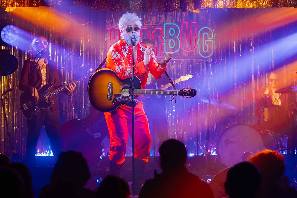 Singer in red suit and sunglasses performs with acoustic guitar on neon-lit stage before crowd, with TRIBUG sign behind.