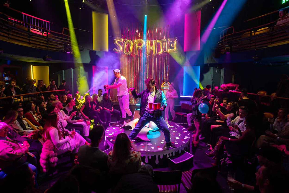 Crowd watches performers on a lit nightclub stage under neon lights, with a gold SOPHIE sign and colorful smoke.