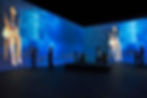People in a dark room watch large blue-lit projections of a woman seated, creating a contemplative and immersive experience.