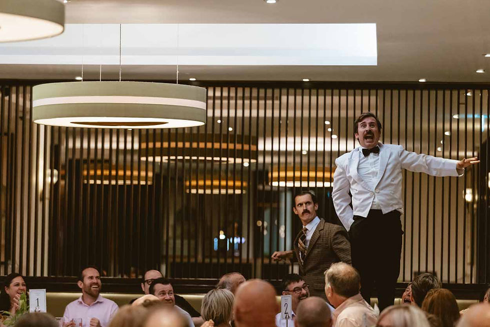 White-jacketed performer stands on a bench at a lively restaurant while laughing guests watch; table numbers 1 and 2 visible.