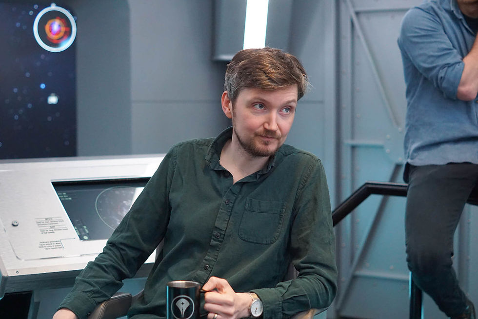 Man in green shirt holding a mug in a futuristic setting. A screen with a circular design is in the background. The mood is contemplative.