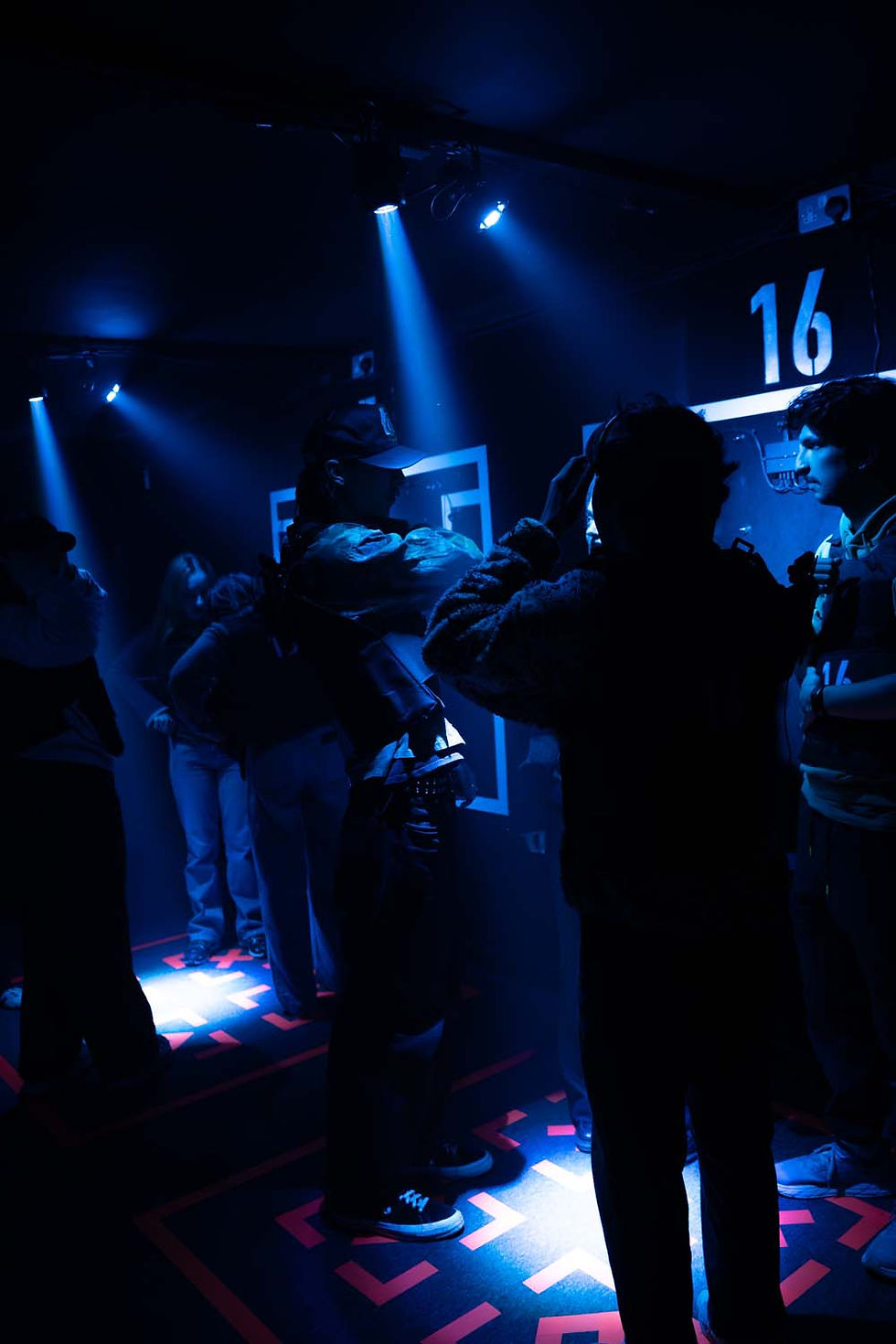 People stand in dim blue-lit room wearing VR headsets, engaging with interactive displays. Floor has red pattern, number 16 on wall.