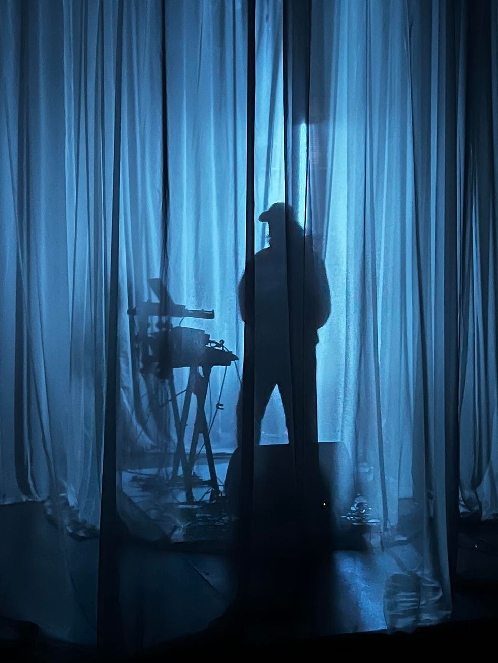 Silhouette of a person with a hat behind sheer blue curtains, standing beside equipment in a dimly lit room, creating a mysterious mood.
