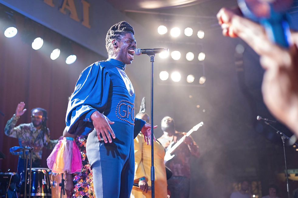 Singer in a blue outfit performs passionately on stage with a band under bright stage lights. Audience arm raised in foreground.