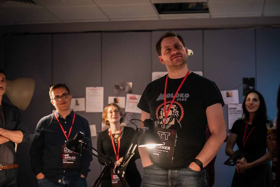 People stand in a dimly lit room with papers on the wall. One person wears a "Moloko Plus" shirt and a badge, looking confident.