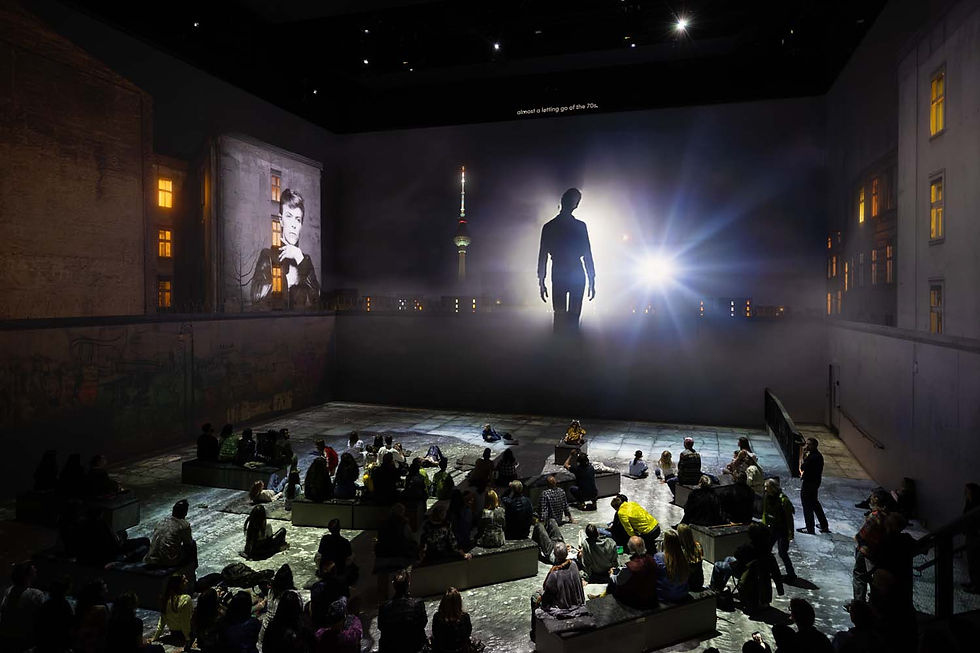 Crowd seated in a dark room watches a large projection. A silhouette and a portrait are visible, with fog effects. Text reads "almost a letting go of the 70s."