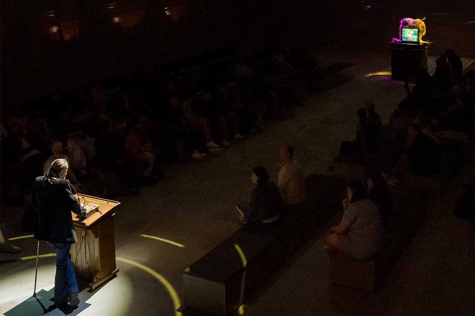 Woman speaks at a podium in a dark auditorium, spotlight on her, with a seated audience and a glowing screen in back.
