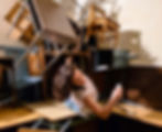 A woman leans back on stacked tables and chairs in a room. She holds paper, surrounded by disarray, creating a chaotic yet calm scene.