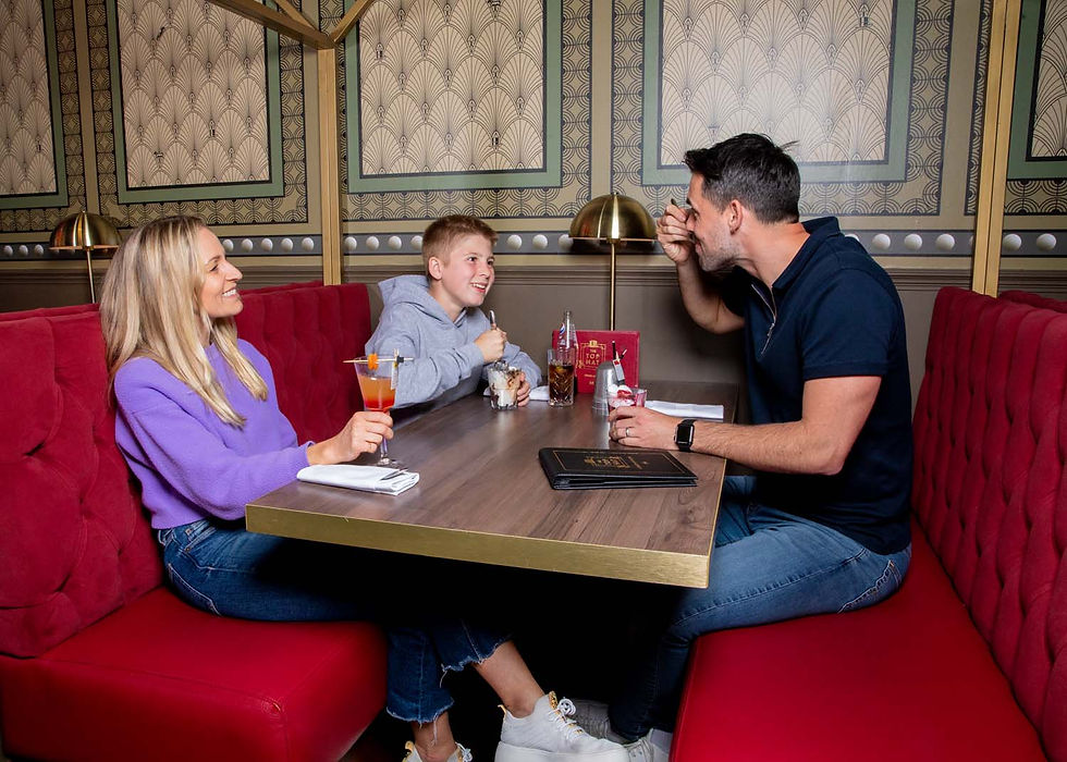 Smiling family of three in a red booth at a restaurant, sharing desserts and drinks at a table with patterned wallpaper behind them.