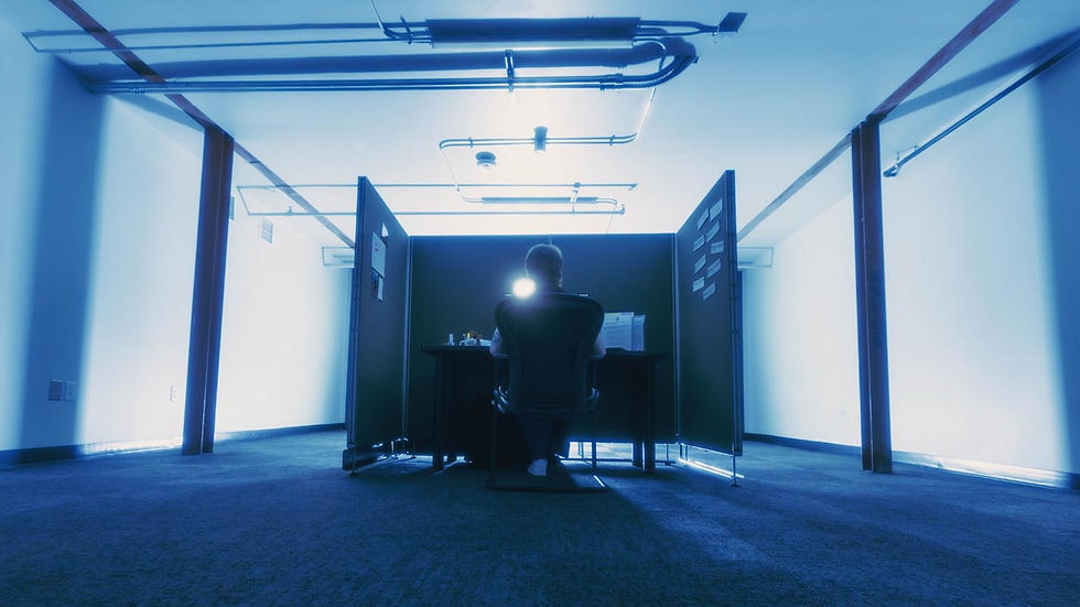 Person sits at a desk in a blue-lit empty office cubicle, seen from behind, with a bright light glowing on the chair.