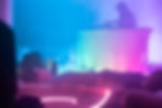 Neon-lit club scene with a DJ at a draped table and a person lying down in the foreground, bathed in blue and pink light.