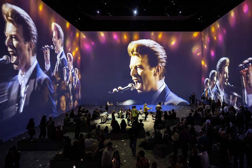 Imposing video projections of a singer performing cover the walls of a dimly lit room. A seated audience watches, creating a vibrant atmosphere.