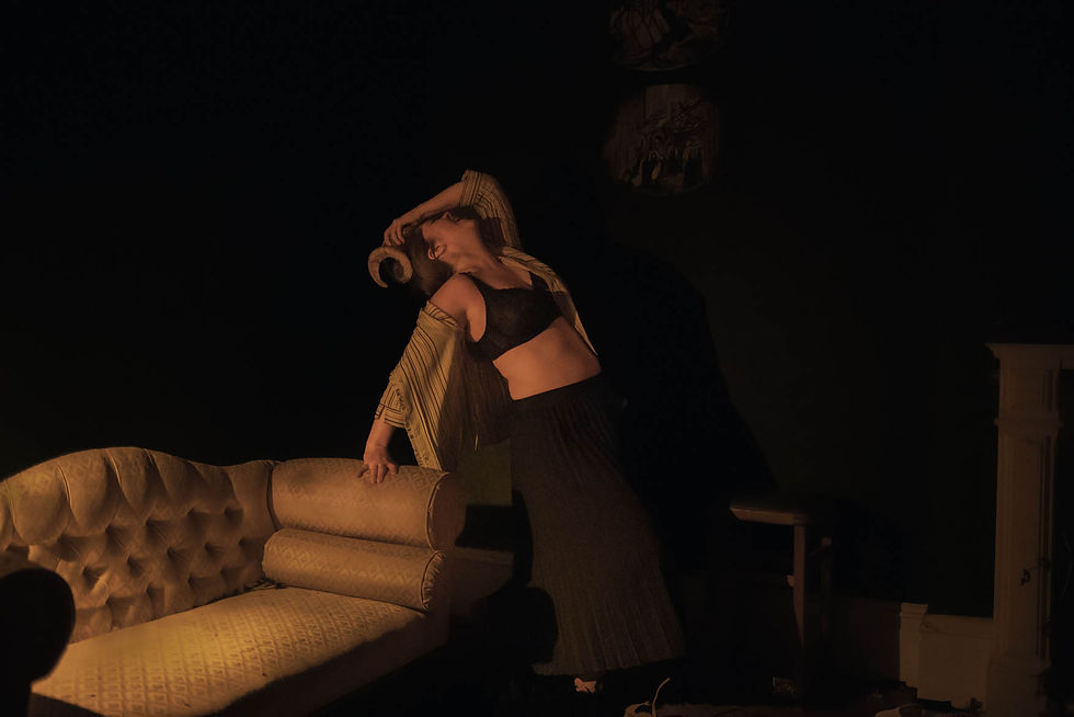 Agatha in dimly lit room wearing a black top and pleated skirt poses on a vintage yellow couch, creating a moody atmosphere.