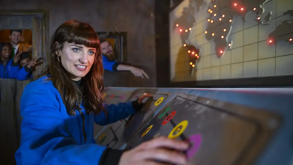 Woman in blue jacket pressing buttons on a control panel, with a world map in the background. Others watch from a doorway, smiling.