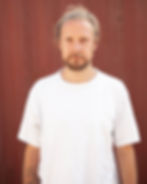 Man with a beard and tied-back hair in a white shirt stands against a red background, displaying a calm expression.