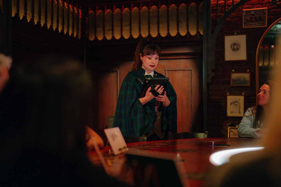 Woman in green plaid cape reads from a tablet in a dim, warm-lit tavern as diners watch.