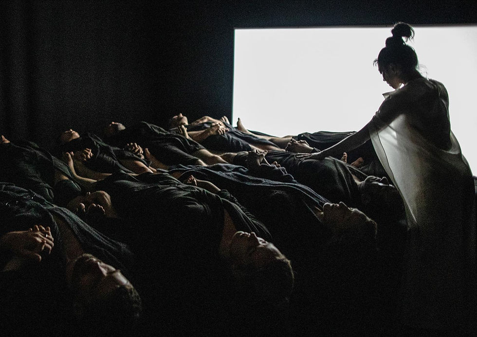 Silhouetted performer in white stands beside many people lying in darkness before a bright screen.