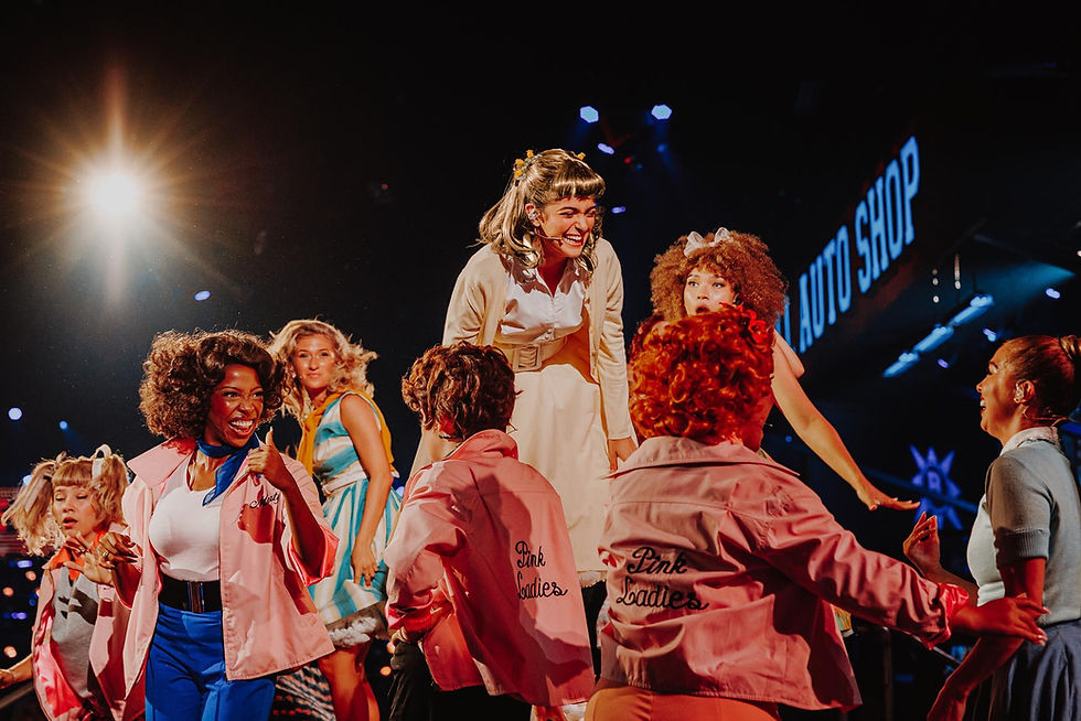 Performers in colorful '50s style outfits, one being lifted, amid vibrant lights. Backdrop reads "Auto Shop," full of energy and joy.